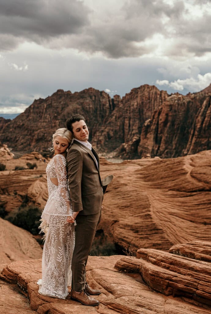 Bride and groom stand back to back on top of rocks with red rocks behind them.