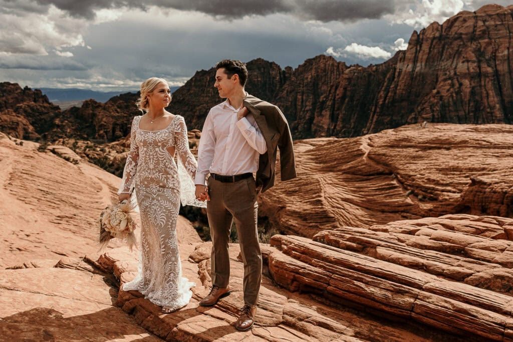 Groom hold jacket over his shoulder as him and the bride look at each other with red rocks in the background.