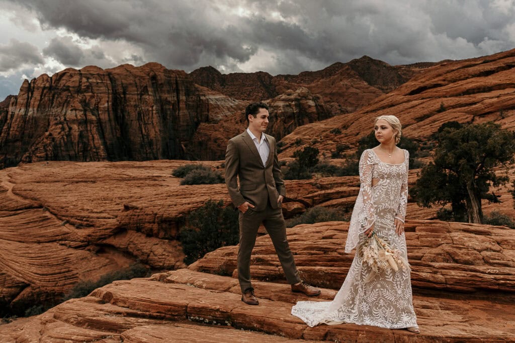 Groom stands behind the bride with red rocks behind them.