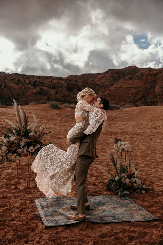 Groom lifts bride during their wedding ceremony on sand dunes.