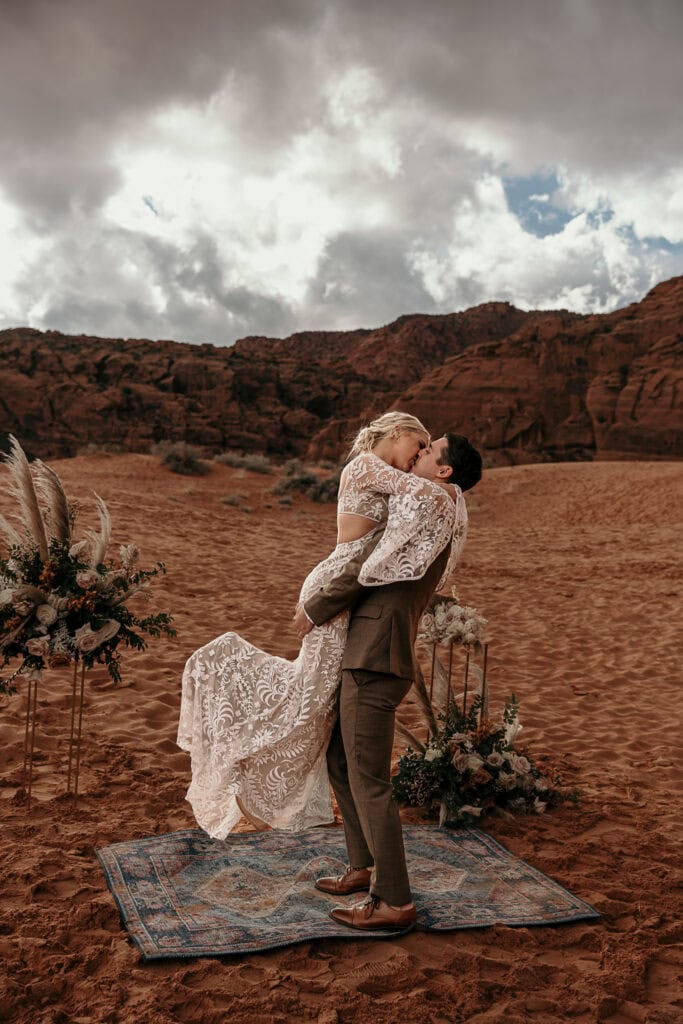 Bride and groom kiss on boho rug on sand dunes.