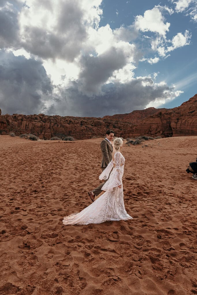 Bride and groom walk on sand dunes in Utah with red rocks and a stormy sky in the background.