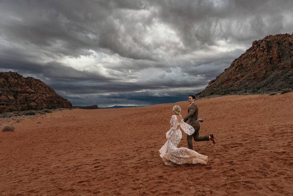 Bride and groom laugh while looking at each other and running on sand dunes during their elopement.