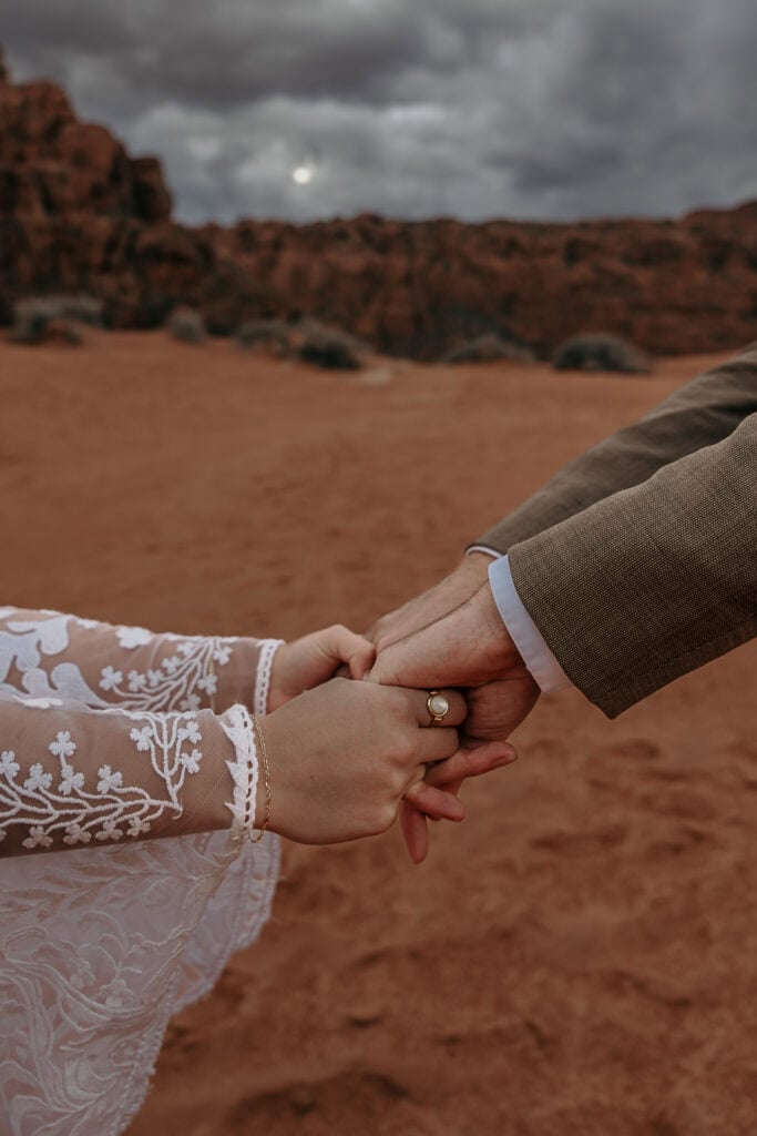 A bride and groom hold hands in front of the red rocks while standing on sand dunes.