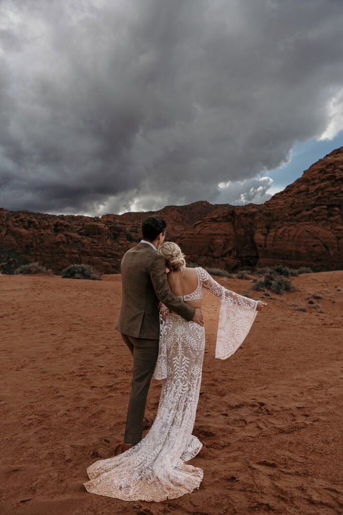 Bride and groom look out over sand dunes and a storm rolling in over red rocks.