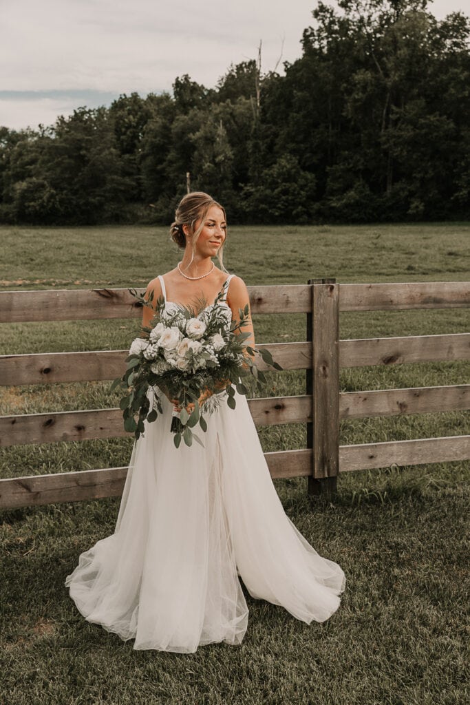 A bride holds her bouquet and looks over her shoulder. A wooden fence is behind her.
