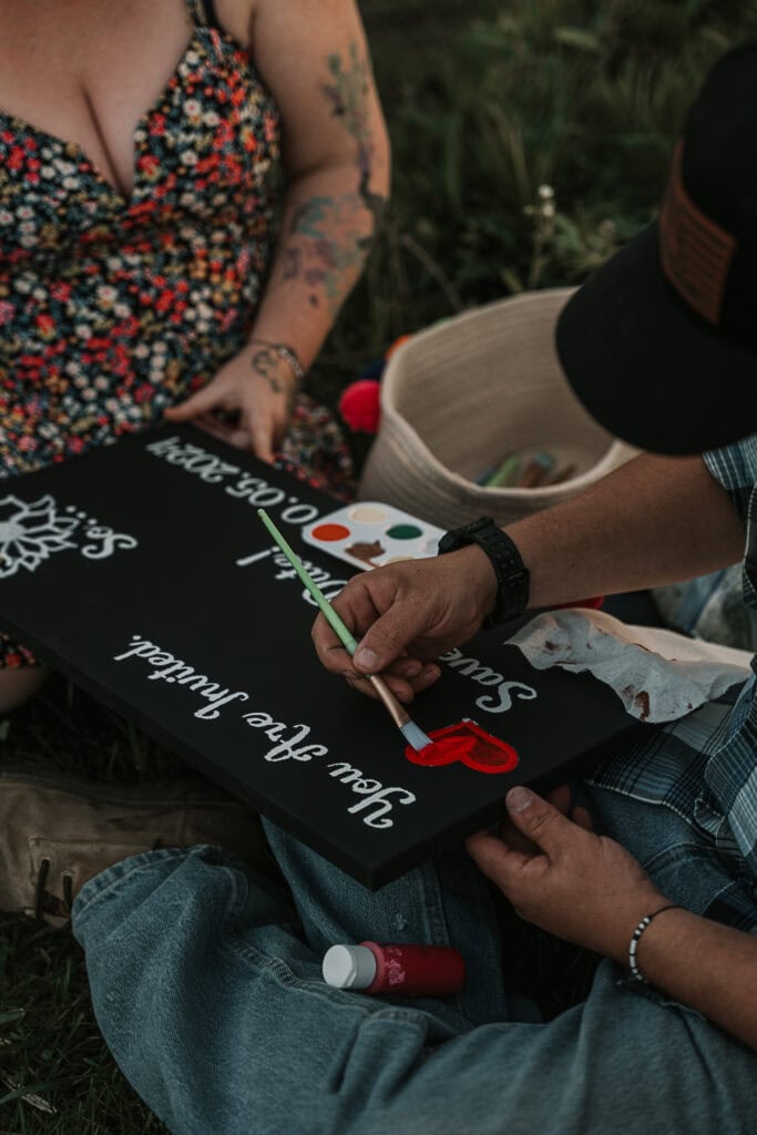 A couple paints a wooden side together while sitting outside.