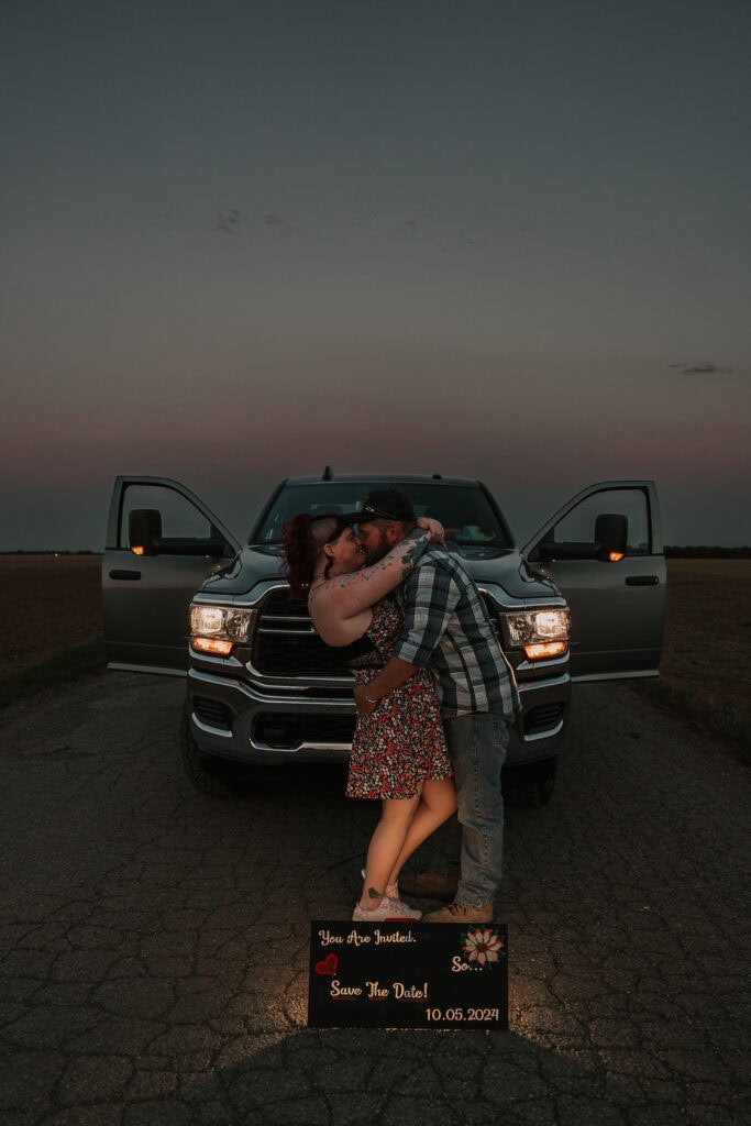 A couple kisses passionately in front of the pickup during sunset. Their save the date is in front of them.