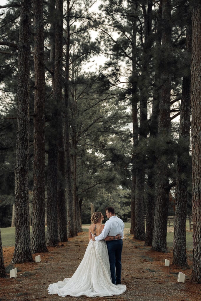 An eloped couple stands in the middle of a row of pine trees with their arms around each other.