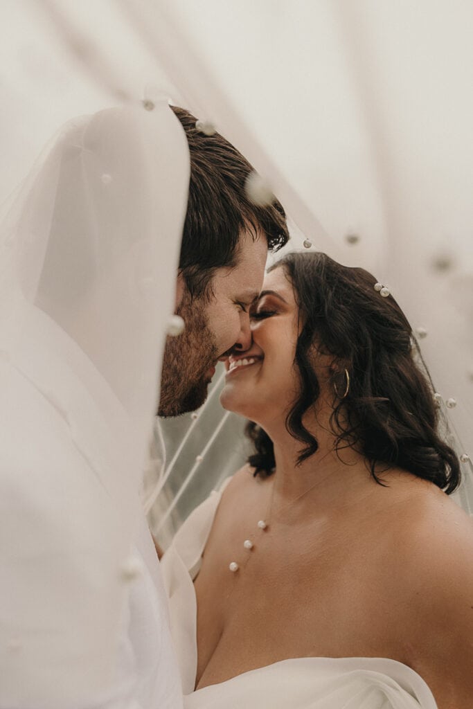 A bride and groom smile while pausing just before a kiss while standing under her pearl detailed bridal veil.