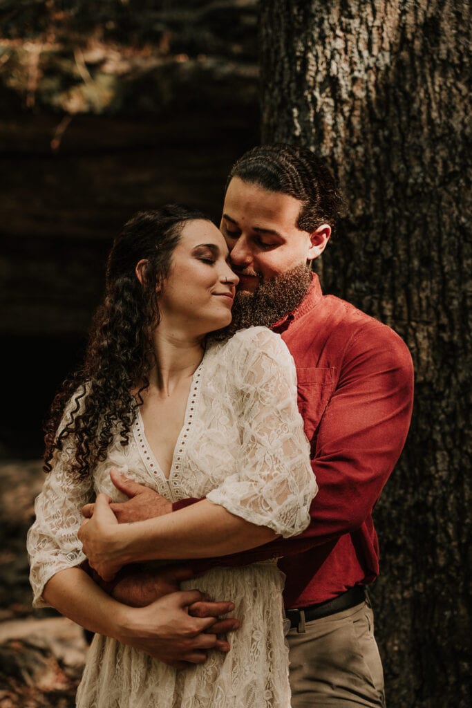 Man leans against a tree and holds his bride around the waist as she leans into him smiling with her eyes closed.
