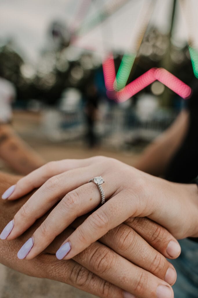 A closeup of a couples hands with hers on the top, showing off her engagement ring. Fair lights are in the background.