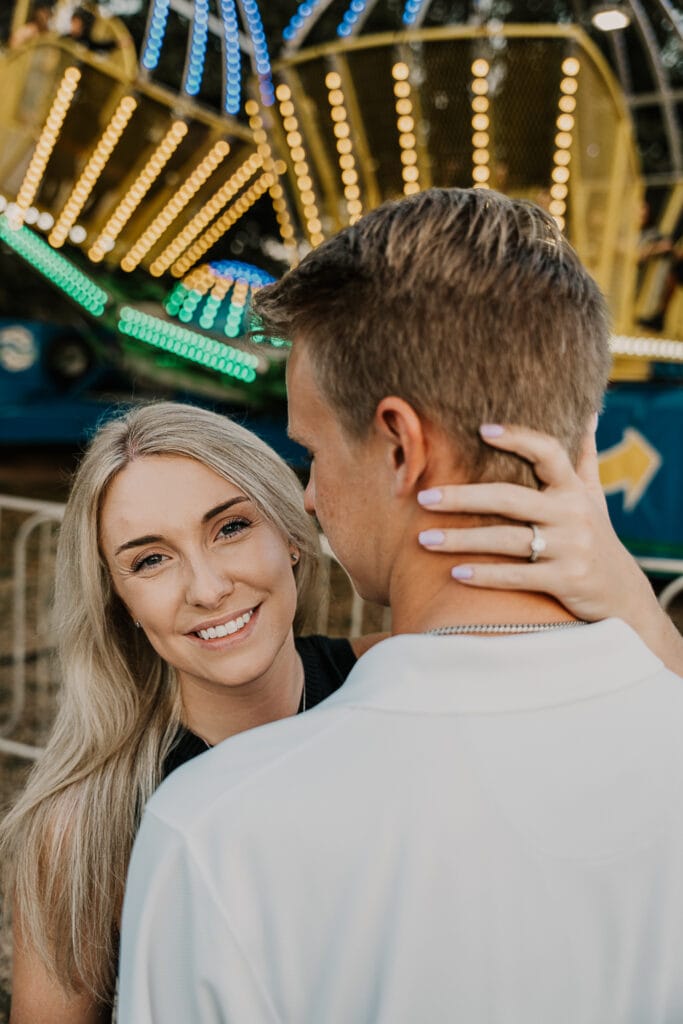 A couple stands facing each other. Her hand is on the back of his head to show off her engagement ring.