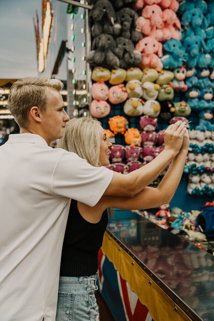 A man shows his fiancee how to throw a dart while they play at the fair.