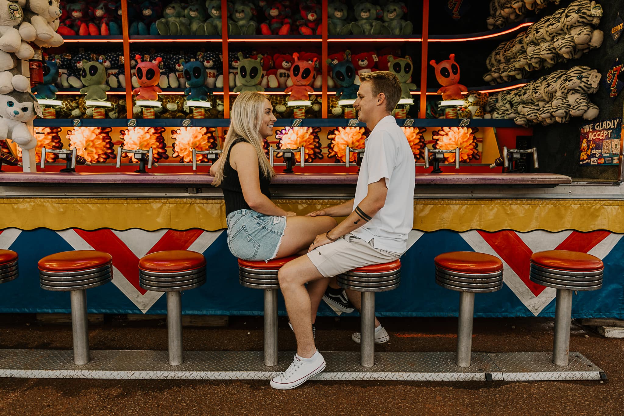 Couple laughs while sitting facing each other in front of a game at the fair.