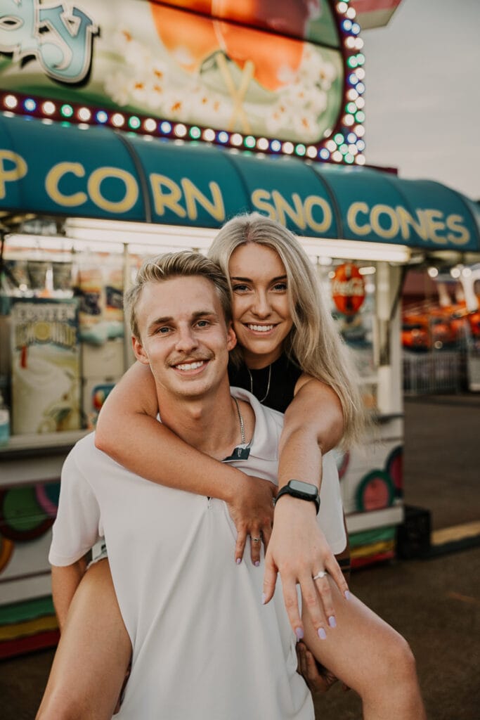 Woman shows off her engagement ring while her fiancé gives her a piggyback ride at the fair.