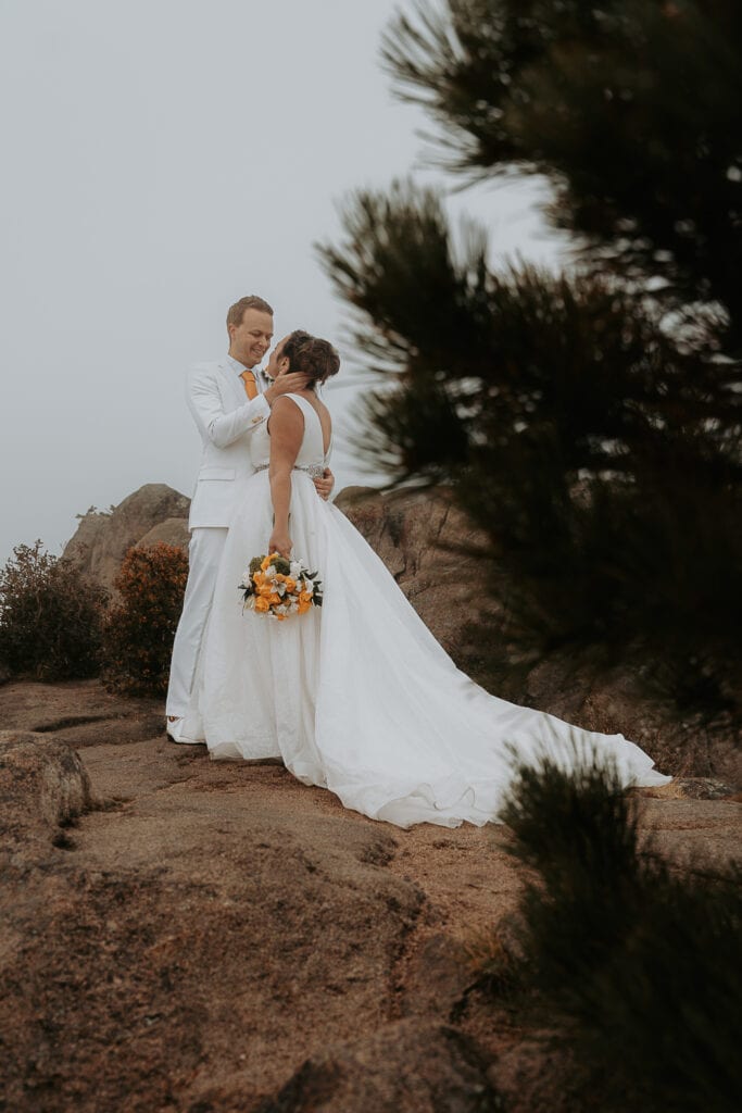 Bride and Groom share a private moment behind a pine tree on top of Kruger Rock in the Rocky Mountains. It is foggy.