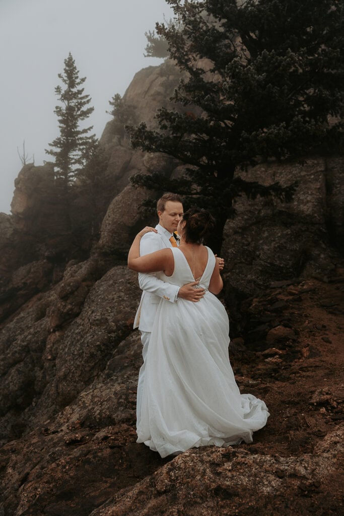 Bride and Groom, both in white, dance in fog on Kruger Rock during Estes Park Colorado elopement.