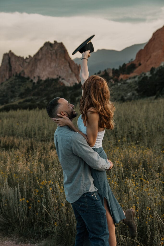Man lifts his girlfriend in the air while she raises her hat. Garden of the Gods sandstone rocks are in the background.