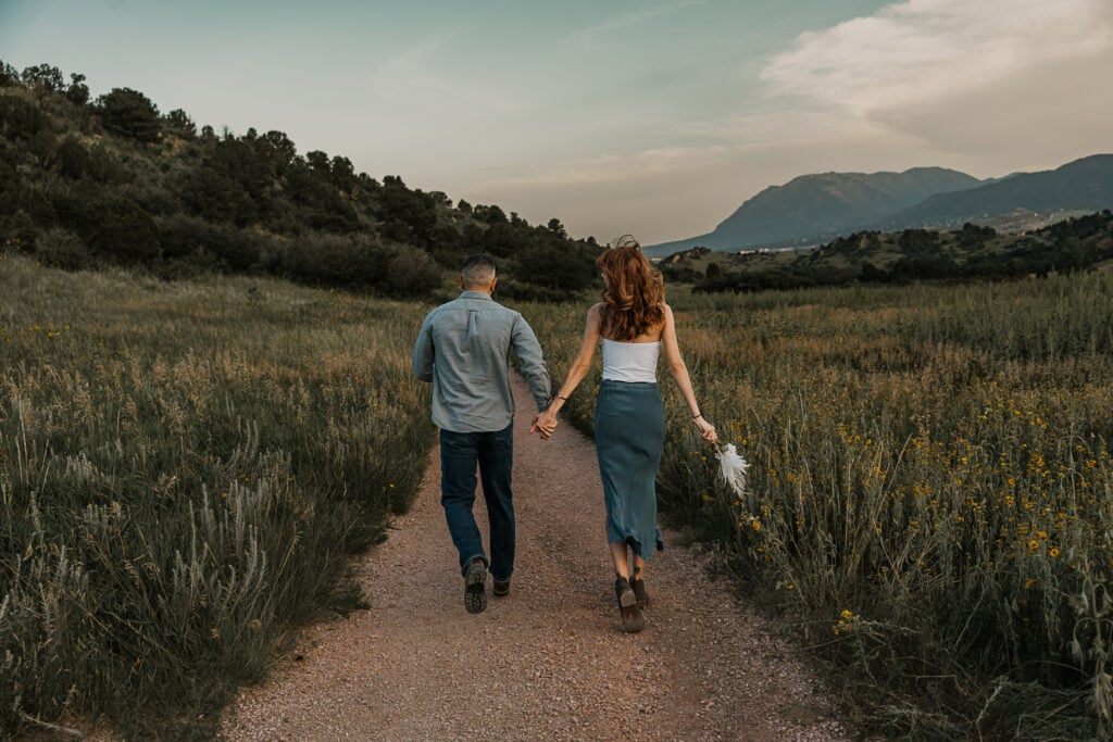 Couple walks hand in hand away from the camera, down a dirt path between wildflowers. A mountain range is in the distance.