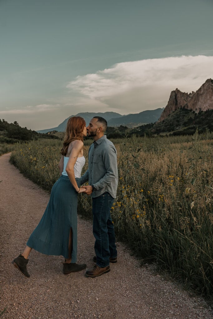 Haley leans in to kiss Charlie while walking along a paved trail in Garden of the Gods. A meadow and flowers are in the background.