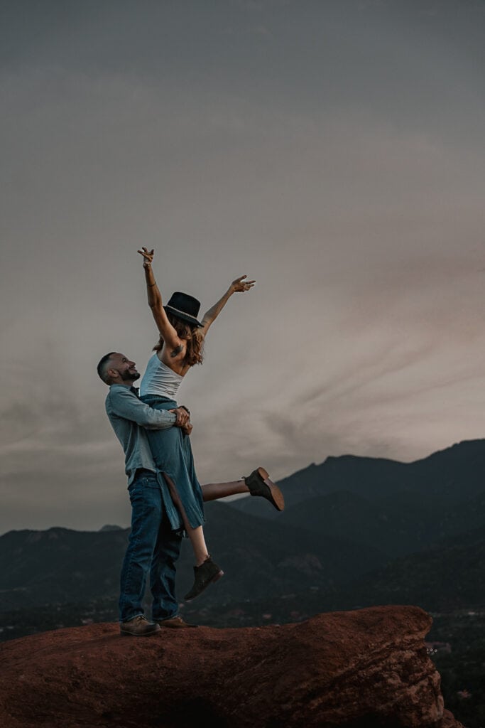 Man standing on a boulder lifts a woman up as she raises her arms in the air during blue hour. The Rocky Mountains are in the background.