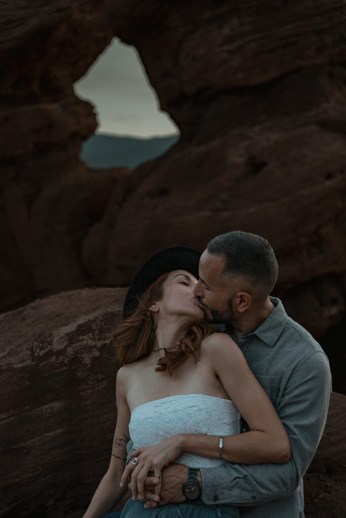 Couple kisses while sitting on rocks. Siamese twins rock formation is behind them.