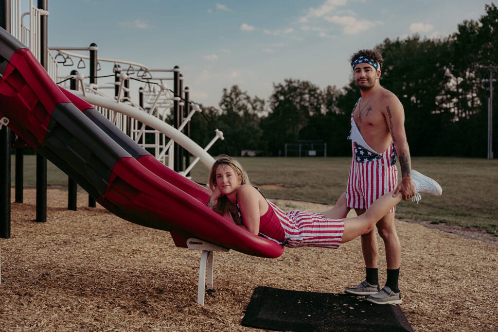 Couple poses for an awkward photoshoot where he holds her feet and she props herself up on a slide in the playground.