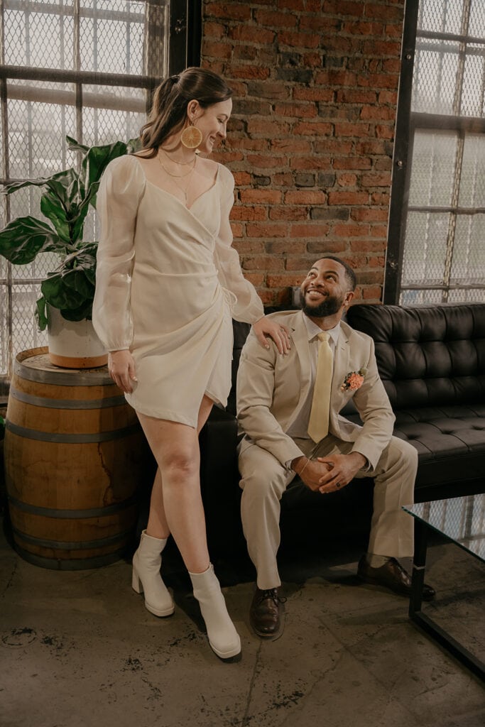 Bride stands with hand on groom's shoulder as he sits on couch inside Corsair Distillery in Nashville during elopement.