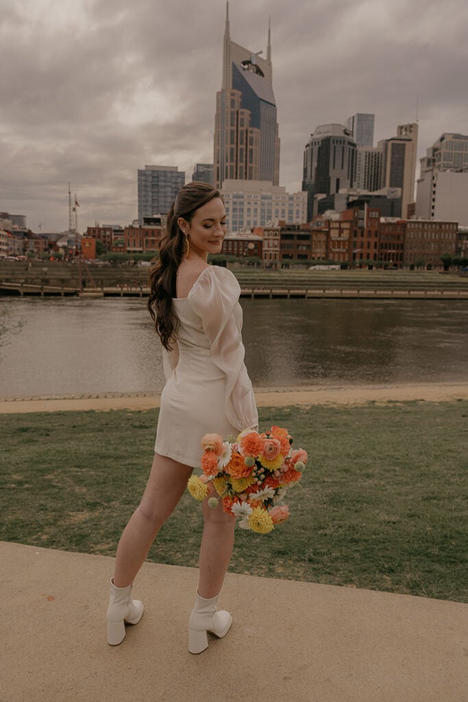 Bride looks down at her citrus bouquet of flowers in Cumberland Park with the Nashville Tennessee skyline behind her