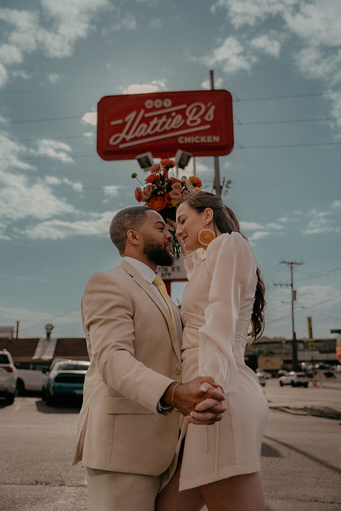 Dakota & Kristin stand facing each other in front of the Hattie B's sign. She is holding her wedding bouquet above them.