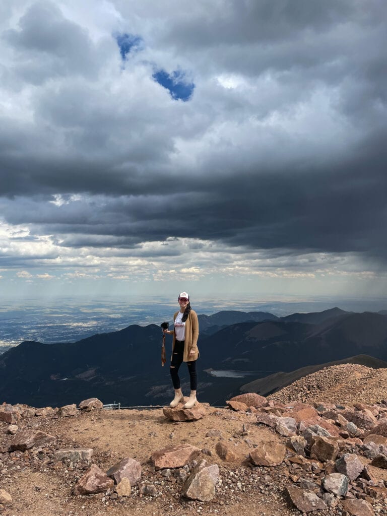 Photographer holds camera in her hand as she stands on a rock on top of Pike's Peak. A storm is coming.