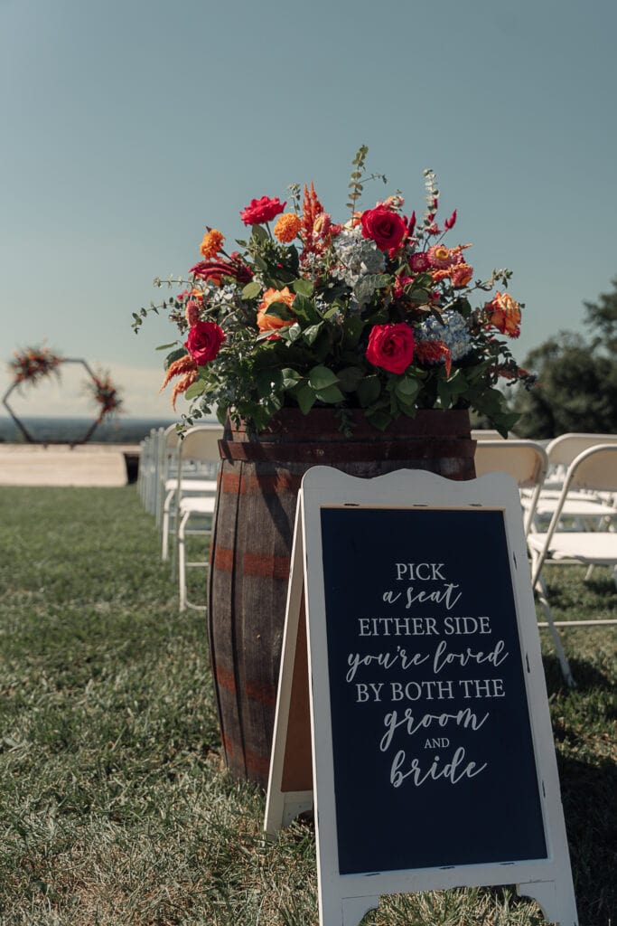 A huge mixed bouquet sits on a barrel at the end of an outdoor wedding aisle. A sign telling people where to sit leans against the barrel. A platform and arch are in the distance.