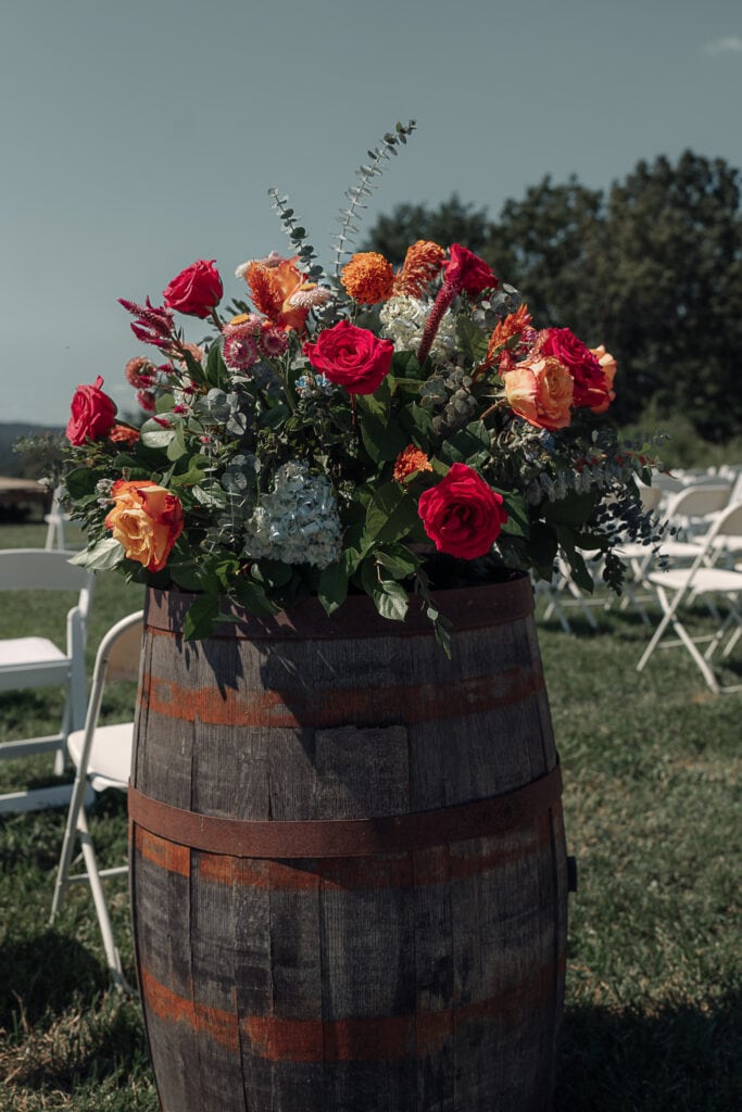A bouquet in a kaleidoscope of colors sets on top of a rustic wooden barrel for an outdoor wedding. While chairs are set up for the ceremony.