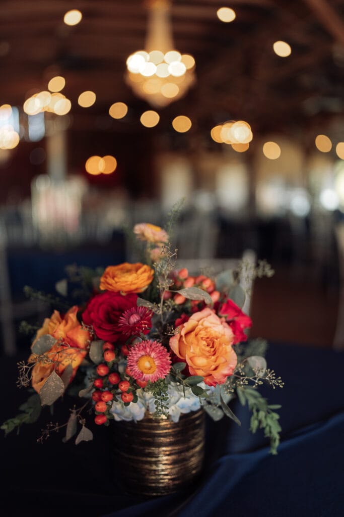 A bouquet of bright mixed flowers sets on top of a navy blue tablecloth in a barn that's set up for a reception. Fairy lights create a bokeh in the background.