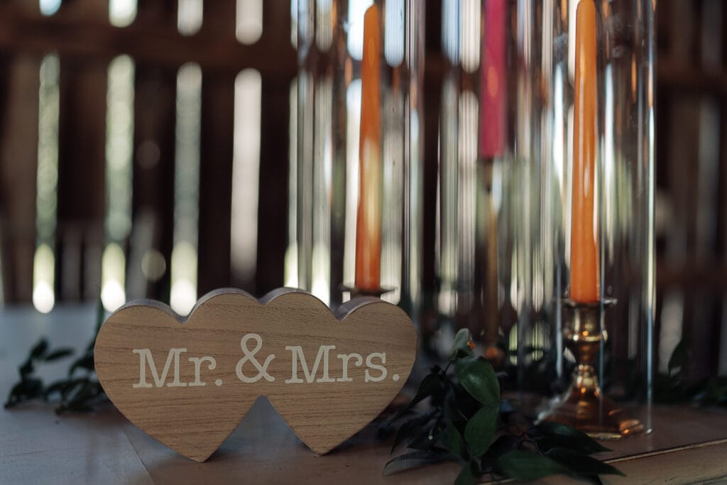 A wooden heart sign that says "MR & MRS" sets on a table at a reception. Orange and pink candlesticks are behind it with greenery.