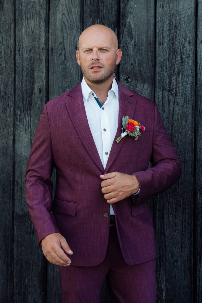 Groom in a plum suit poses with one hand on the button of his jacket.