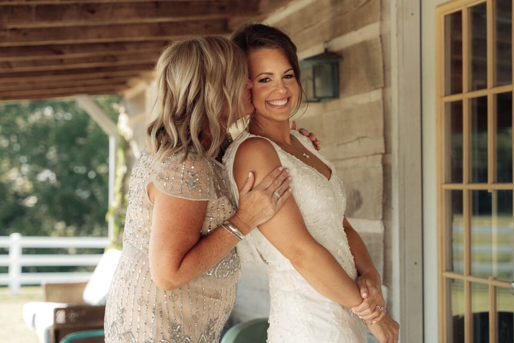 The mother of the bride pulls her daughter in from behind her shoulders as she kisses her cheek.