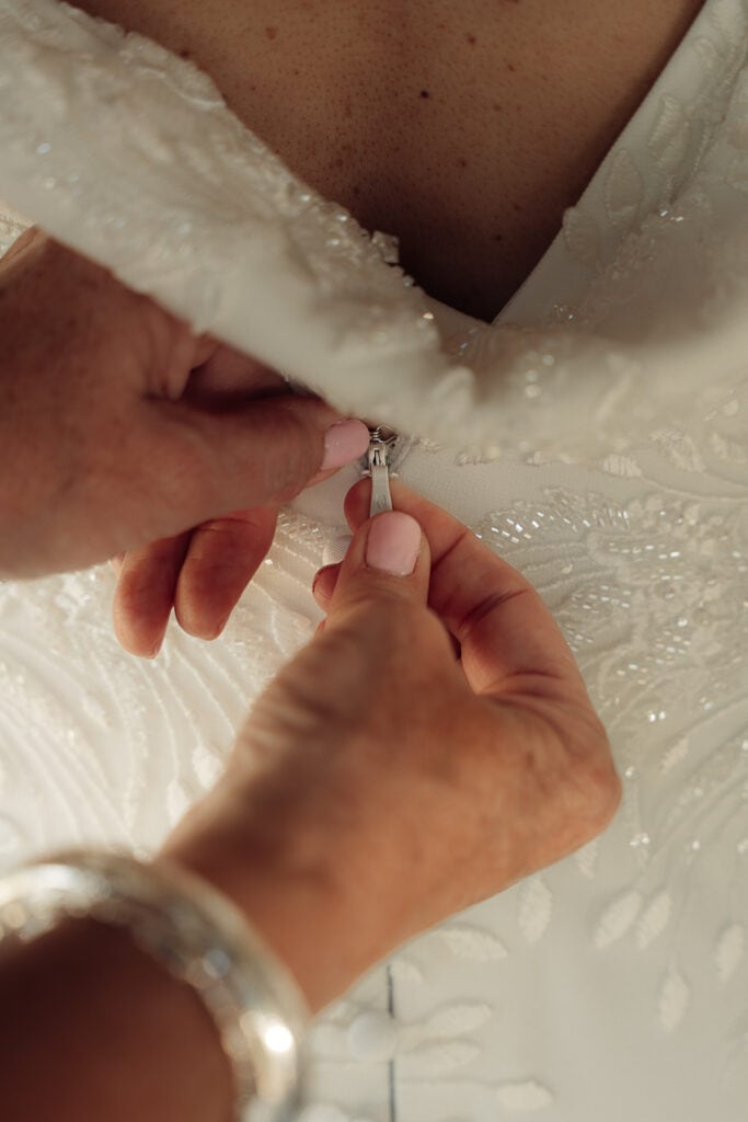 A closeup image of a bride's mom's hands zipping up her wedding gown.