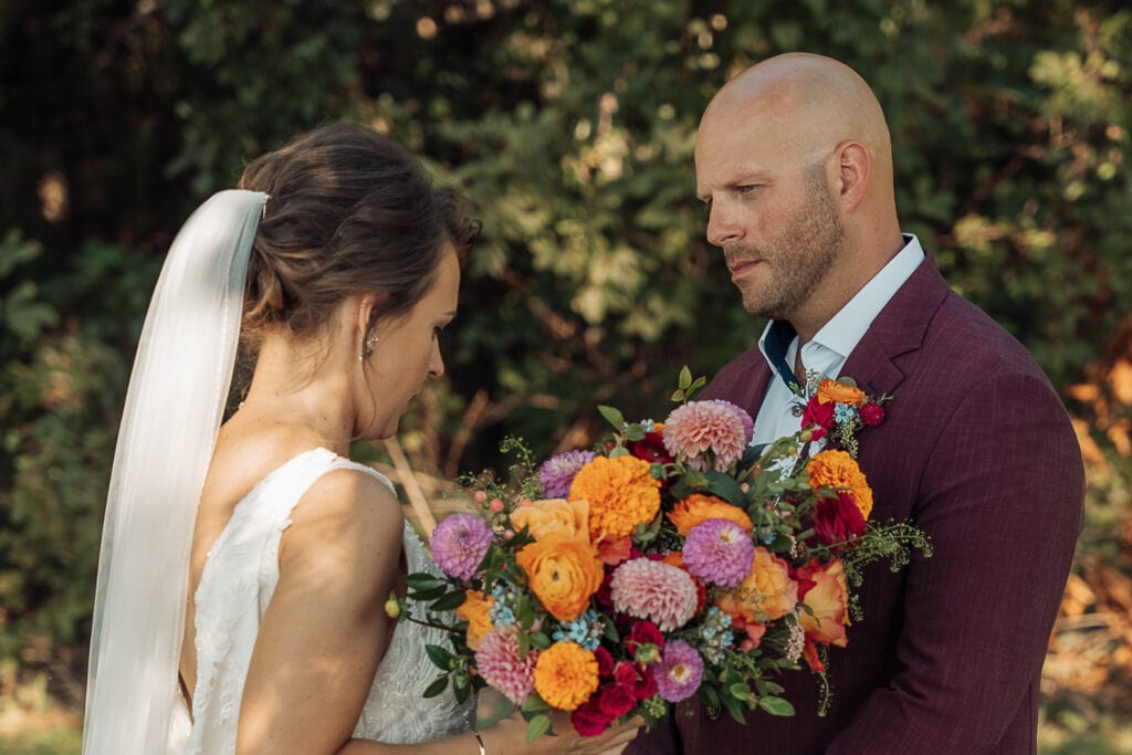 A groom looks intently at the Bride as she reads something to him during their first look on their wedding day. She is holding a giant bouquet of bright flowers.
