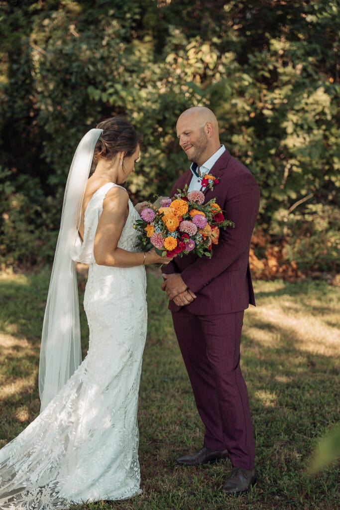 AJ smiles at Lauren with his hands folded as she stands in front of him, reading something to him before their wedding. She is holding a giant bouquet of vibrant flowers.