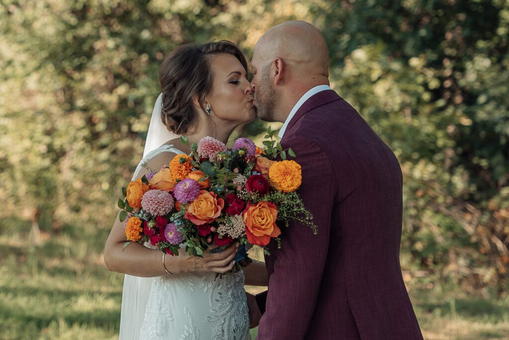 A Bride and Groom kiss while standing outside under the shade of a tree. She is holding a big vibrant bouquet.