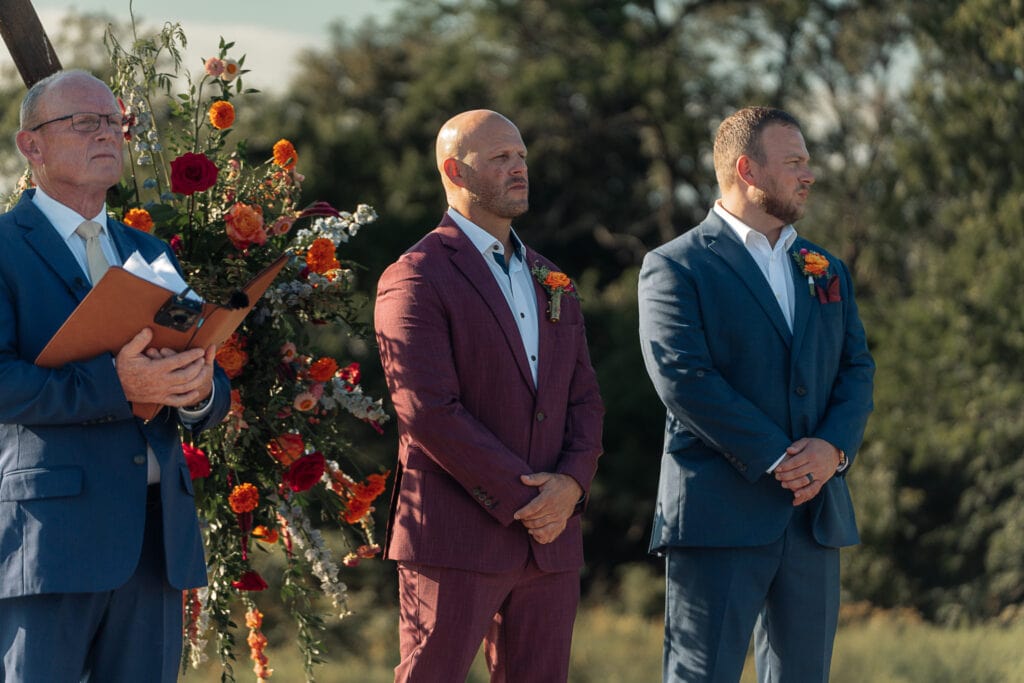 A groom stands at the altar waiting for the bride to walk down the aisle at their outdoor wedding. The officiant and best man stand on each side of him.