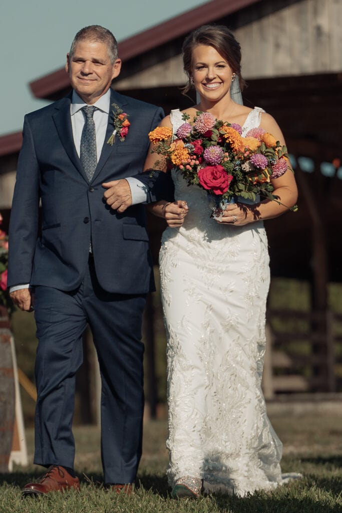 A father walks his daughter down the aisle during an outdoor wedding ceremony. A barn is in the background.