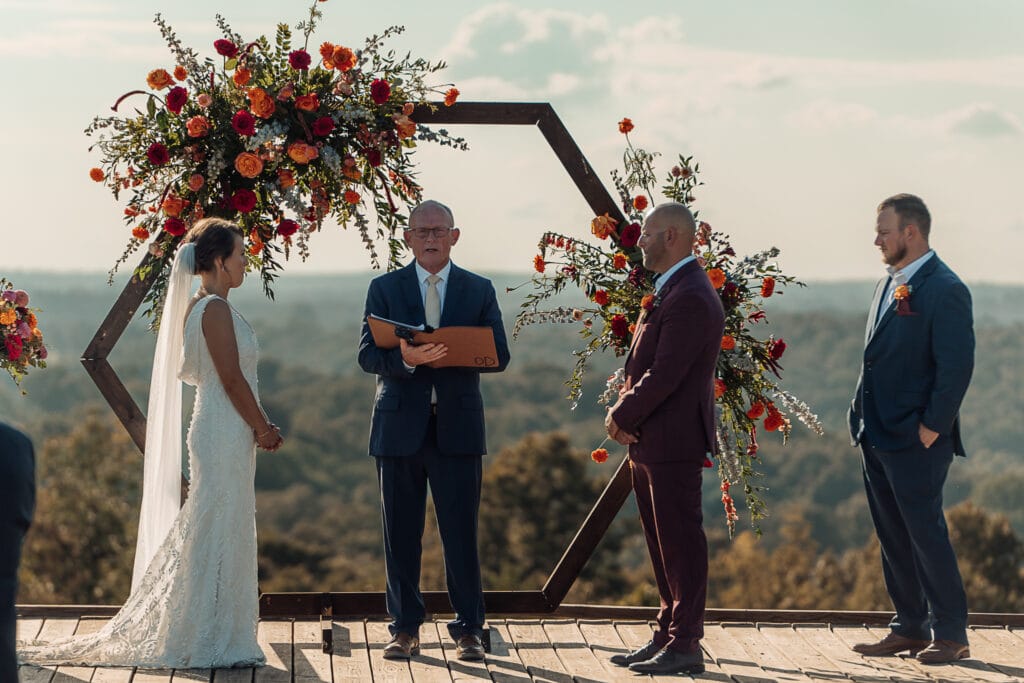 A man officiates an outdoor wedding as the wedding party stands on a wooden platform in front of a floral decorated wooden arch. Rolling hills are in the background.