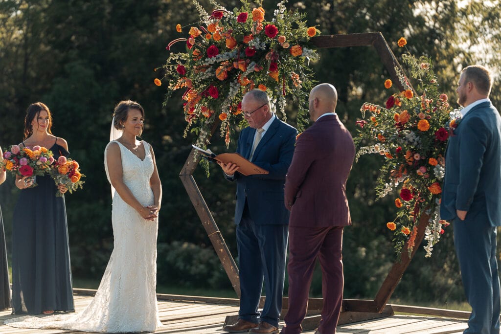 A bride and groom smile at each other while a man officiates their outdoor wedding. The wedding party stands on a platform with them. A wooden arch with flowers is behind the officiant.