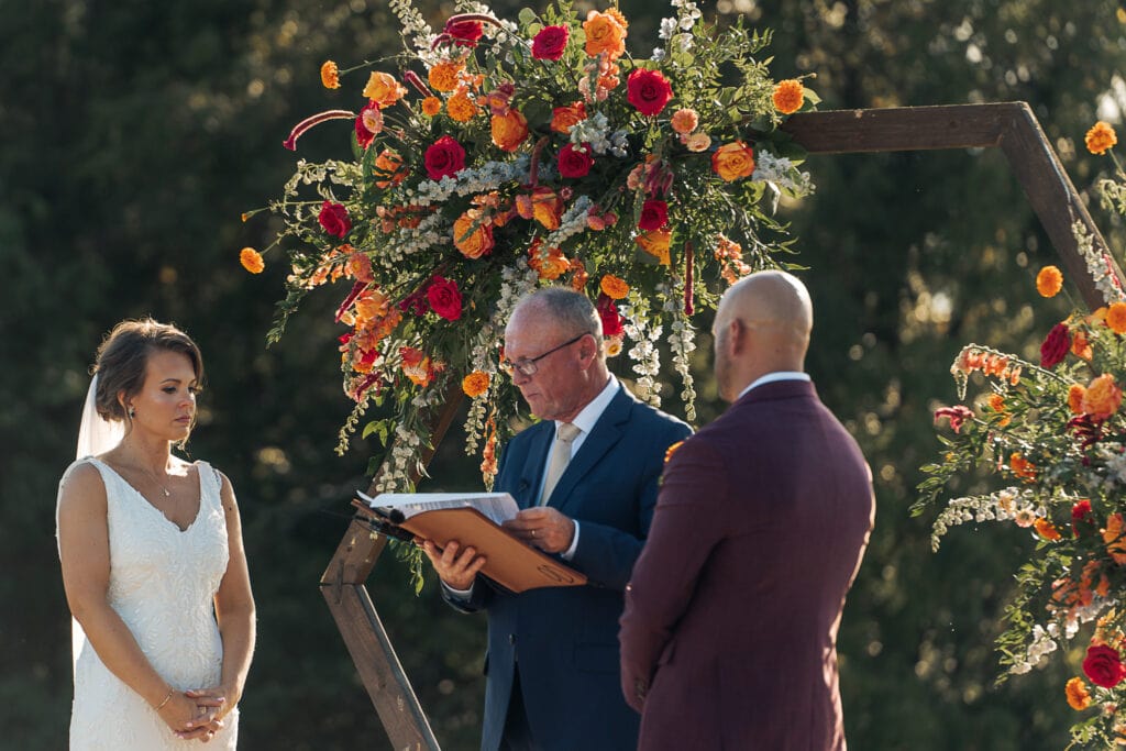 An officiant reads from his binder during an outdoor wedding ceremony. The sun shines on the massive bouquets decorating the arch behind him and the bride and groom.