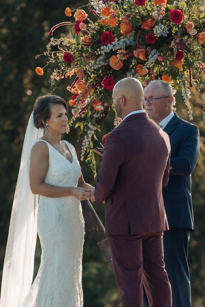 Bride & Groom exchange vows in front of an arch that's decorated with vibrant flowers.