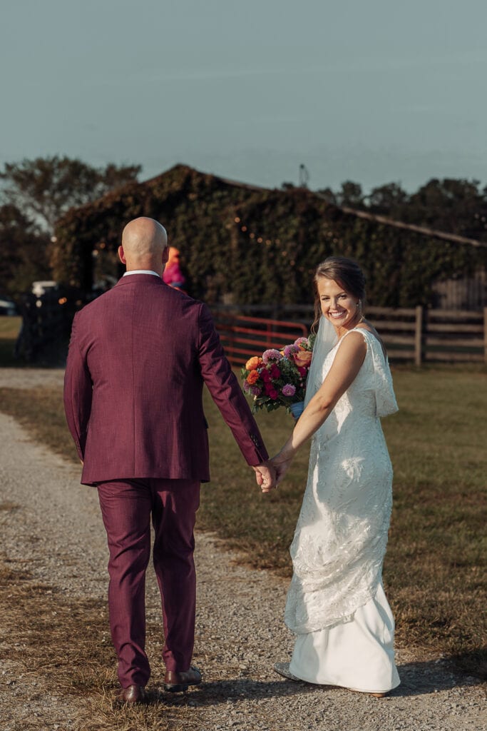 Bride & Groom are walking towards a barn, holding hands. The Bride looks back at the camera.