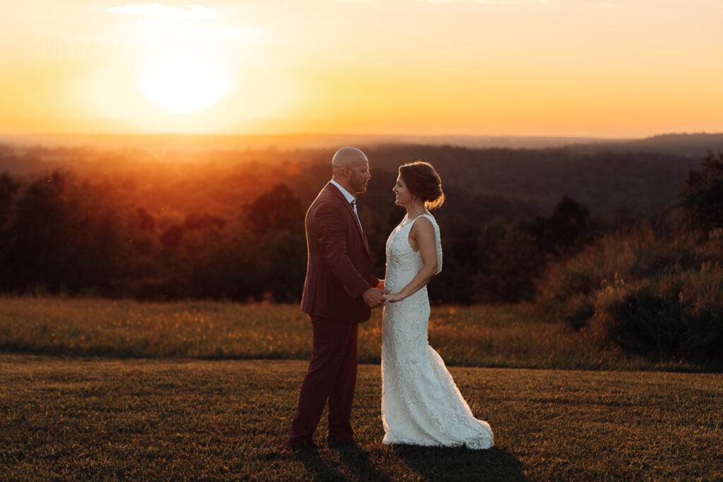 Man and woman hold hands while looking at each other during sunset. They're wearing wedding attire.